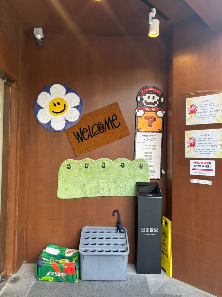 매장 입구 벽면에 꾸며진 소품들입니다. 우드톤 벽면에 꽃 모양 장식과 WELCOME 문구, 그리고 픽셀 느낌의 캐릭터 장식이 있어서,