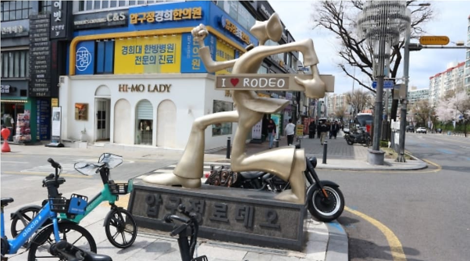 압구정 로데오역 인근에 세워진 '압구정 로데오' 조형물과, 그 옆 골목의 화려한 매장 건물입니다. 이 거리의 이름과 지금 얼굴을 함께 보여줍니다.