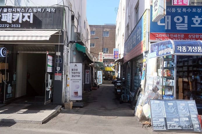 간판이 빼곡히 걸린 좁은 골목과, 벽면 가득 물건이 쌓인 안쪽 통로입니다. 좁아지는 보행 폭을 실감할 수 있는 장면입니다.