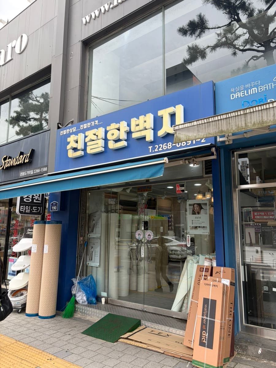 벽지 도매 가게 정면과, 그 옆에 쌓인 벽지 샘플 상자입니다. 개인 셀프 인테리어 수요가 몰리는 대표 업종을 보여줍니다.