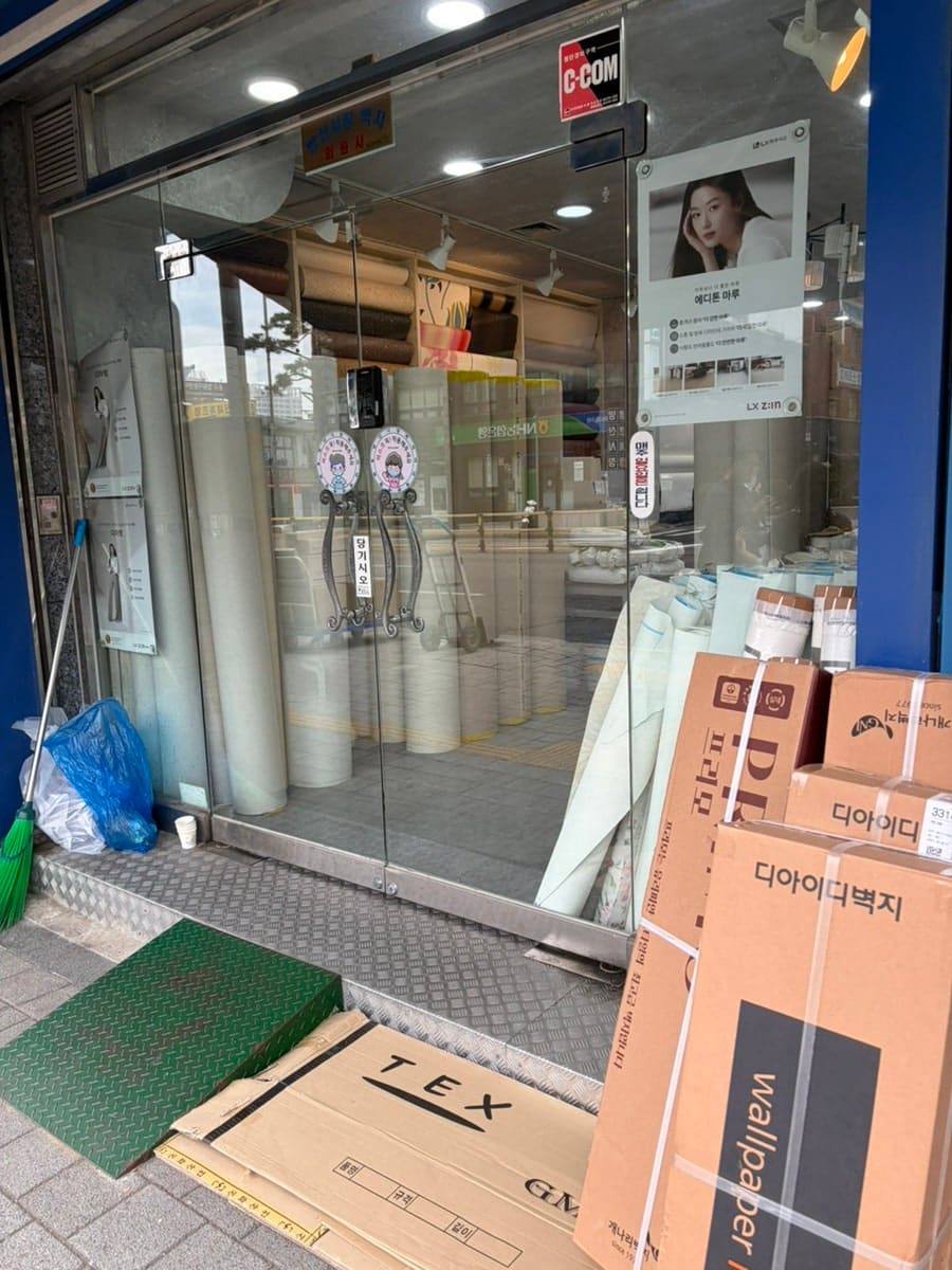 벽지 도매 가게 정면과, 그 옆에 쌓인 벽지 샘플 상자입니다. 개인 셀프 인테리어 수요가 몰리는 대표 업종을 보여줍니다.