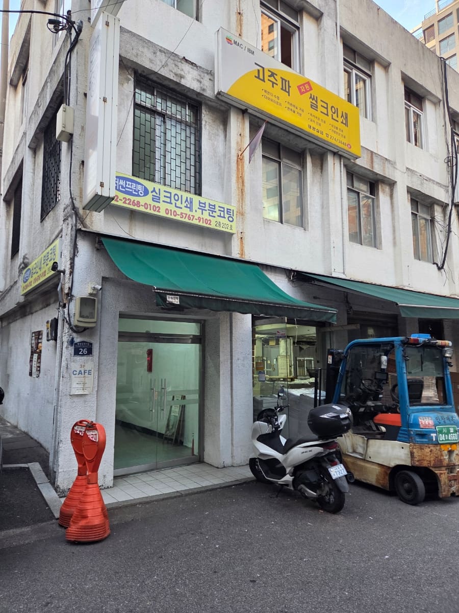 같은 사람이 조금 멀리서 찍은 건물 전경입니다. '고주파 씰크인쇄' 노란 간판과 포크레인, 오토바이가 세워진 인쇄소 골목의 평범한 한 장면입니다.