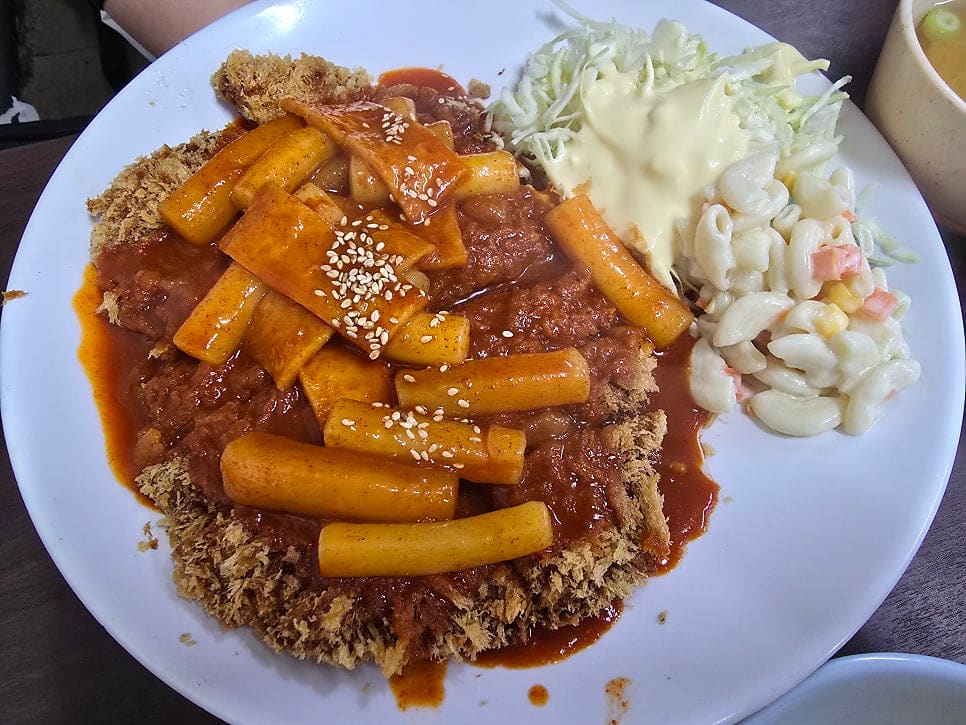 같은 사람이 가까이서 찍은 돈까스+떡볶이 사진입니다. 참깨가 뿌려진 떡볶이 소스가 튀김옷 위를 가득 덮고 있습니다.