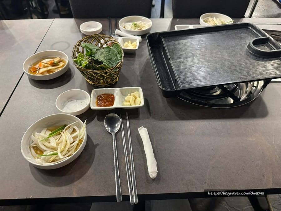 같은 사람이 주문 직후 찍은 상차림 사진입니다. 상추와 깻잎이 담긴 바구니, 양파절임과 쌈장, 마늘이 아직 불을 올리지 않은 불판 옆에 놓여 있습니다.