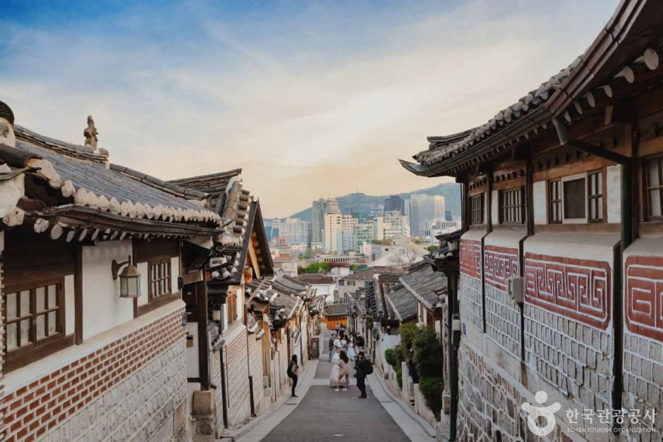왼쪽은 낮 시간대 계동길 골목. 한옥 기와지붕 사이 좁은 통로를 관광객들이 걷고 있고 멀리 도심 빌딩이 겹쳐 보인다. 오른쪽은 해질 무렵 같은 골목으로,