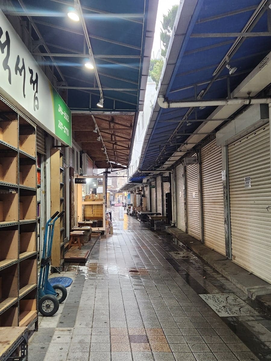 가족사진이 걸린 책방 외벽과, 비에 젖은 채 셔터가 늘어선 골목 통로다. 사람이 없는 아침, 젖은 바닥 위로 정적이 내려앉는다.