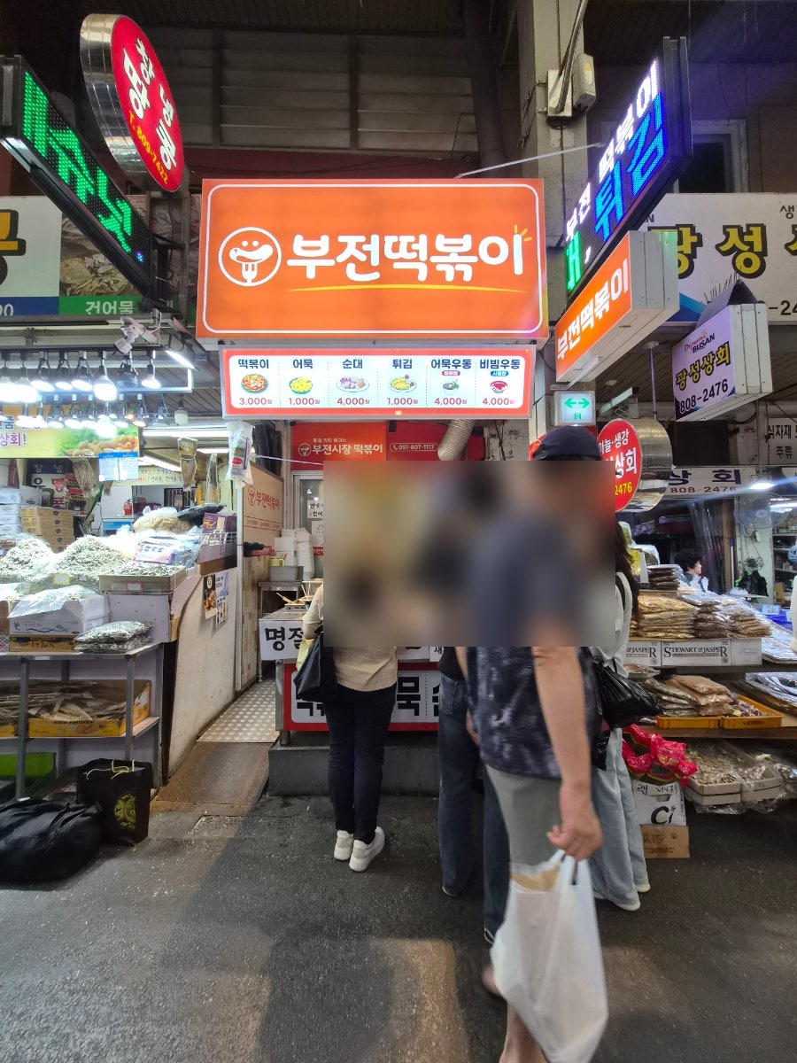 suhyun5618이 담은 부전떡볶이 좌판입니다. 주황 간판 아래 사람들이 서 있고, 다른 한 장에는 검은 장갑을 낀 손이 봉투에 음식을 담아 건네는 순간이 찍혀 있습니다.