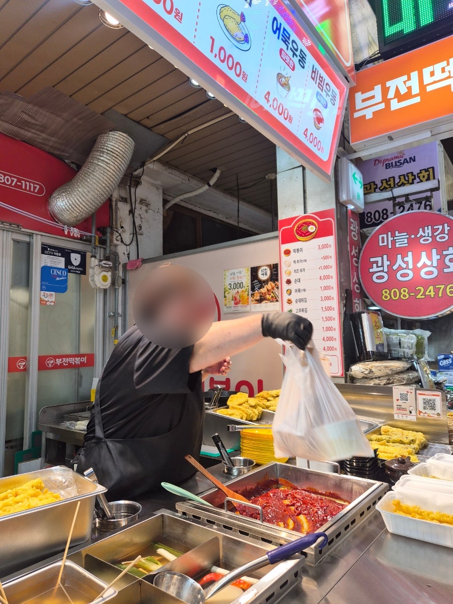 suhyun5618이 담은 부전떡볶이 좌판입니다. 주황 간판 아래 사람들이 서 있고, 다른 한 장에는 검은 장갑을 낀 손이 봉투에 음식을 담아 건네는 순간이 찍혀 있습니다.