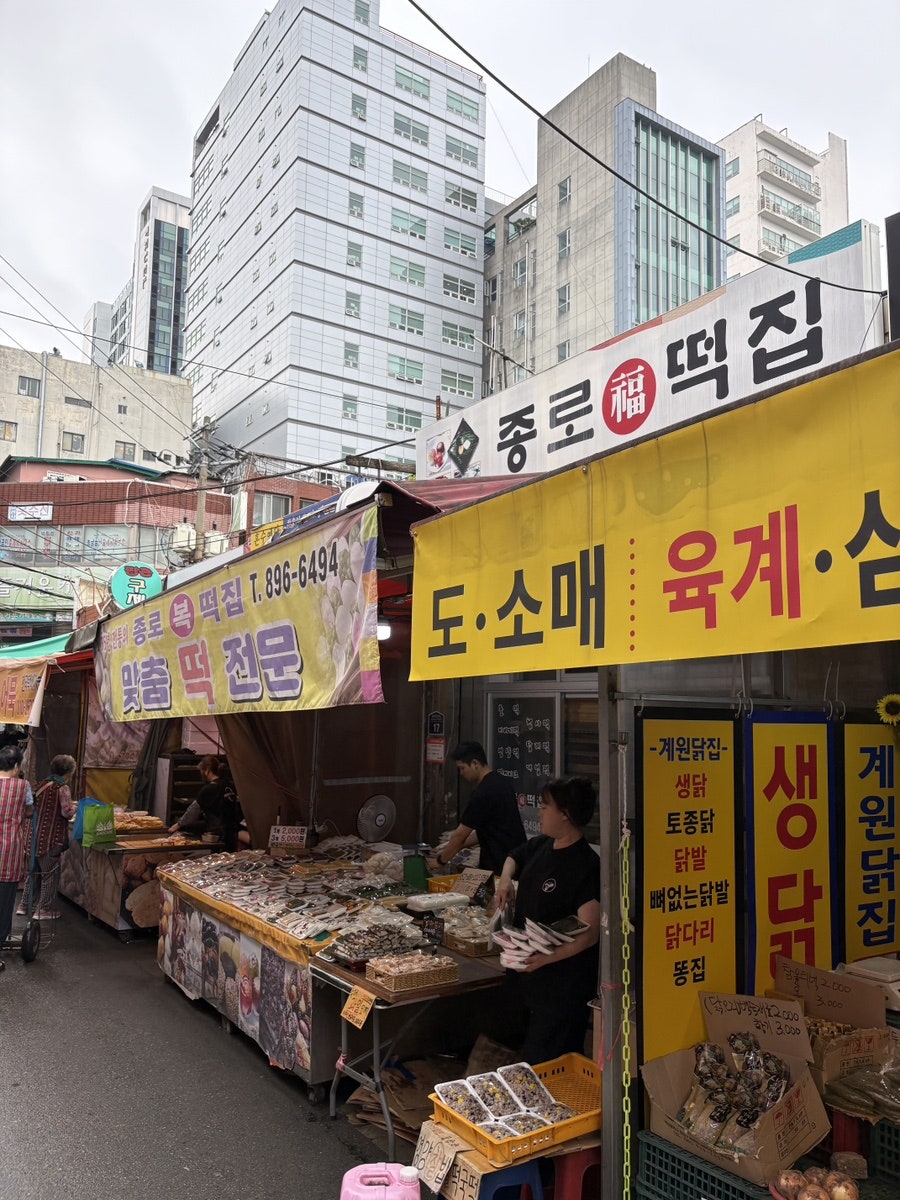 duoying332가 지붕 없는 바깥 골목에서 담은 사진입니다. '종로福떡집'과 생닭집 차양이 나란히 걸린 좌판 뒤로 높은 빌딩들이 올려다보이고,