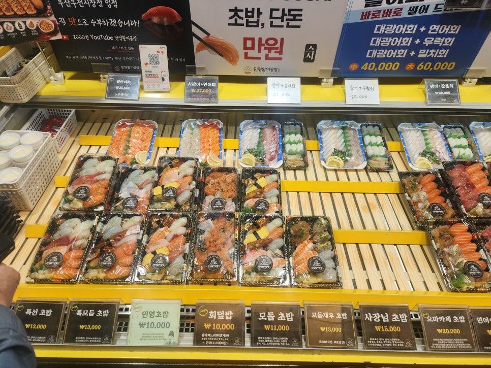 leesunyou가 저녁 무렵 담은 사진입니다. 불을 환하게 밝힌 가게 전경과, 광어·연어·우럭·모둠초밥이 가격표와 함께 진열대 가득 늘어선 모습이 나란합니다.