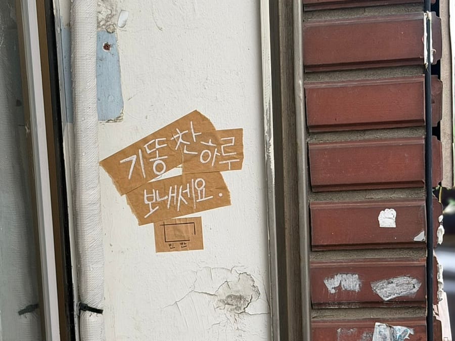 카페 입구 옆 낡은 벽에 손글씨로 적힌 쪽지를 발견한 블로거는 그 장면을 가까이서 찍어 남겼습니다. 낡은 벽돌과 벗겨진 페인트, 그 위에 붙은 '기똥찬 하루 보내세요'라는 글씨,