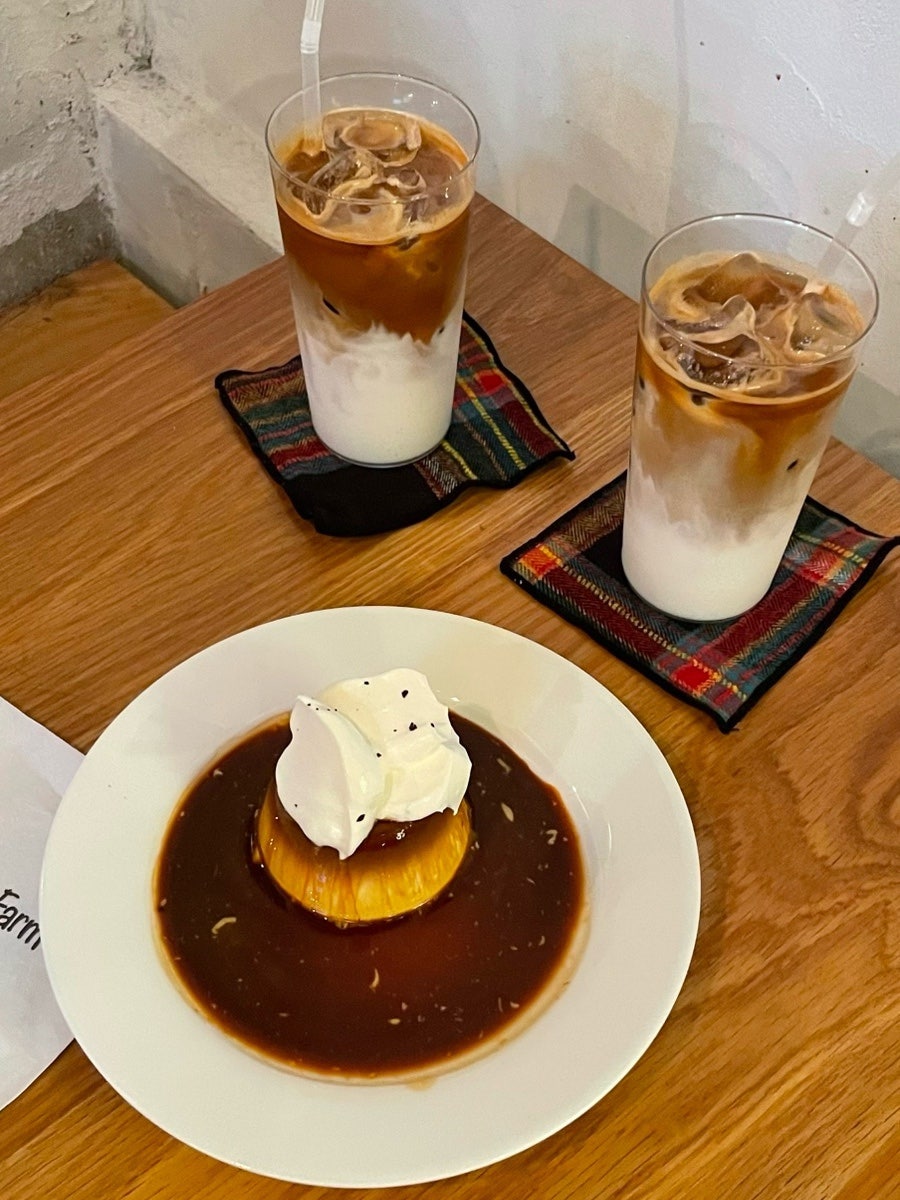 bongz__가 담은 장면입니다. 옷걸이 행거와 책이 놓인 벽감이 있는 넓은 실내, 그리고 체크무늬 코스터 위 아이스 라떼 두 잔과 캐러멜크림푸딩을 가까이서 찍었습니다.