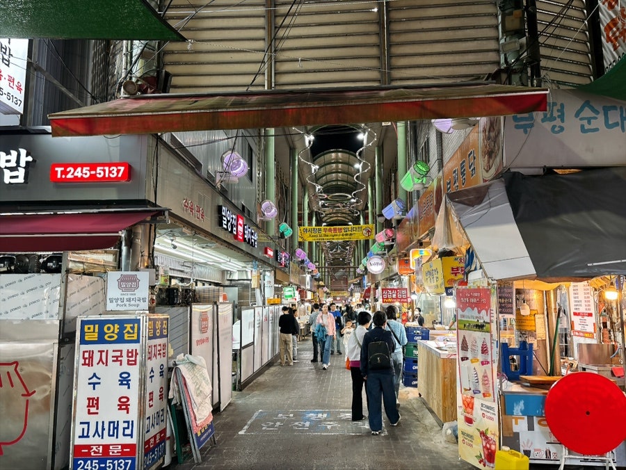 밤이 되어 조명이 켜진 깡통시장 통로입니다. 알전구와 포장마차 매대 사이로 사람들이 줄지어 걷고, 2013년부터 운영된 국내 1호 야시장이라는 안내문도 통로 한편에 서 있습니다.
