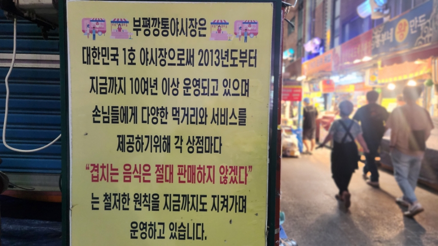 밤이 되어 조명이 켜진 깡통시장 통로입니다. 알전구와 포장마차 매대 사이로 사람들이 줄지어 걷고, 2013년부터 운영된 국내 1호 야시장이라는 안내문도 통로 한편에 서 있습니다.
