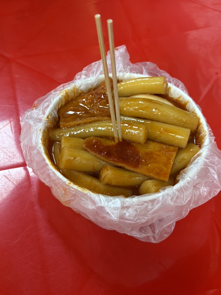 비닐을 씌운 그릇에 담긴 떡볶이가 빨간 테이블 위에 놓여 있습니다. 유리문에 붙은 노란 종이들마다 다녀간 사람들의 손글씨 메시지가 적혀 있습니다.