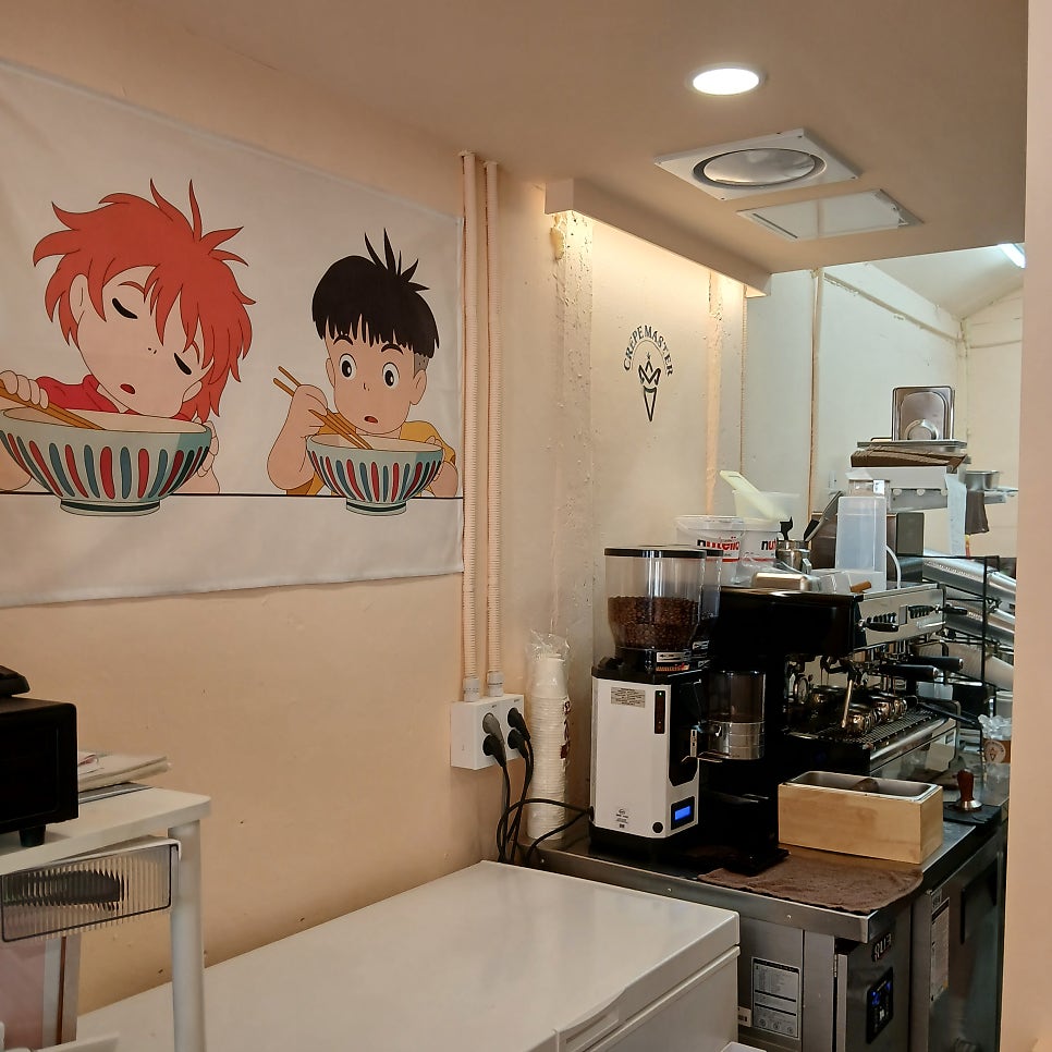 CRÊPE MASTER 간판이 처마 아래 걸려 있고, 좁은 내부에는 에스프레소 머신과 함께 포뇨 캐릭터가 그려진 천이 걸려 있습니다.