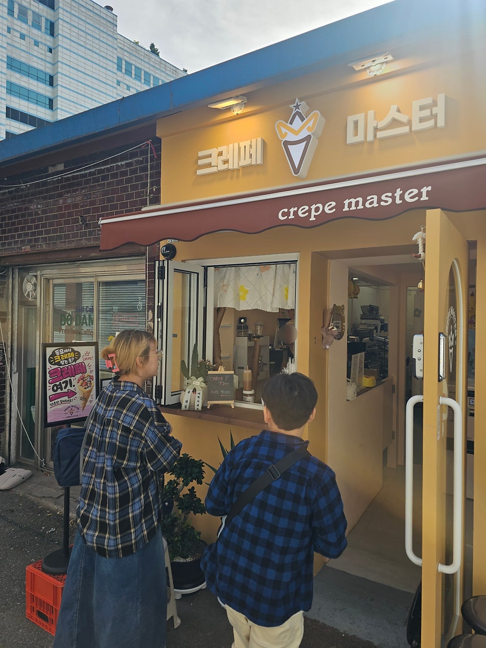 좁은 골목 끝자락, 노란 외벽의 크레페마스터가 보입니다. 창 너머로 주문을 넣는 손님들의 뒷모습도 함께인데,