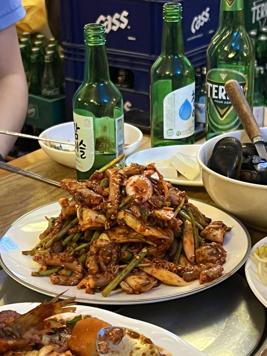 초록 소주병과 맥주병 사이로 매콤하게 무친 해산물 요리가 수북이 쌓여 있고, 갓 구워 낸 감자전을 사장님이 직접 테이블로 가져다주고 있습니다.