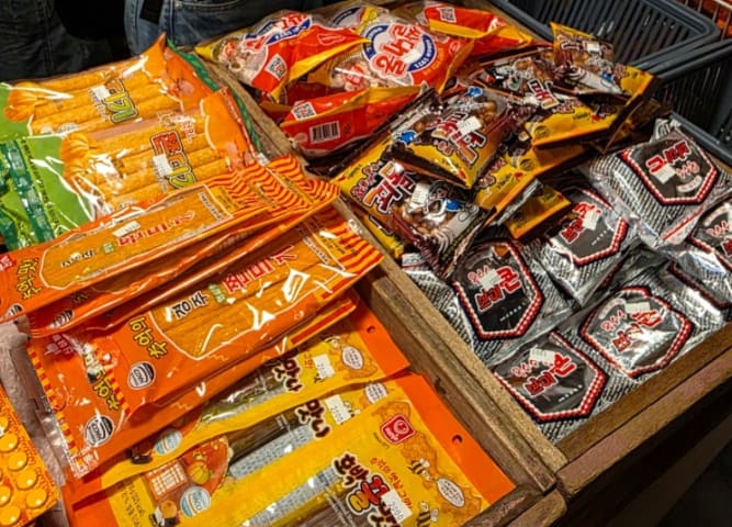 천장까지 빼곡한 옛날 장난감 사이로 사람들이 줄지어 구경합니다. 계산대 옆엔 알록달록한 옛날 과자 봉지들이 상자째 쌓여 있어,