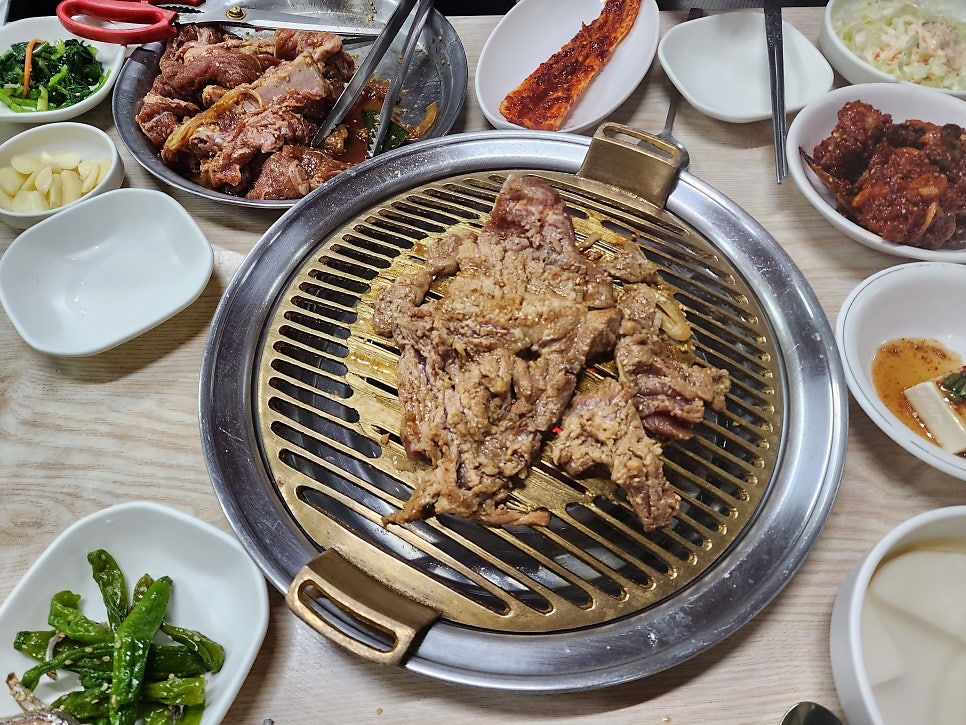 화로를 중심으로 상추쌈과 양념게장, 콩나물 같은 반찬이 가득 둘러싸고 있고, 노릇하게 구워진 돼지갈비가 불판 위에서 지글거립니다. 낡은 간판과 좁은 길,