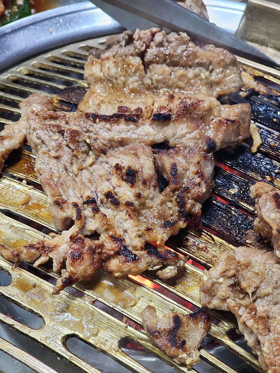 화로를 중심으로 상추쌈과 양념게장, 콩나물 같은 반찬이 가득 둘러싸고 있고, 노릇하게 구워진 돼지갈비가 불판 위에서 지글거립니다. 낡은 간판과 좁은 길,