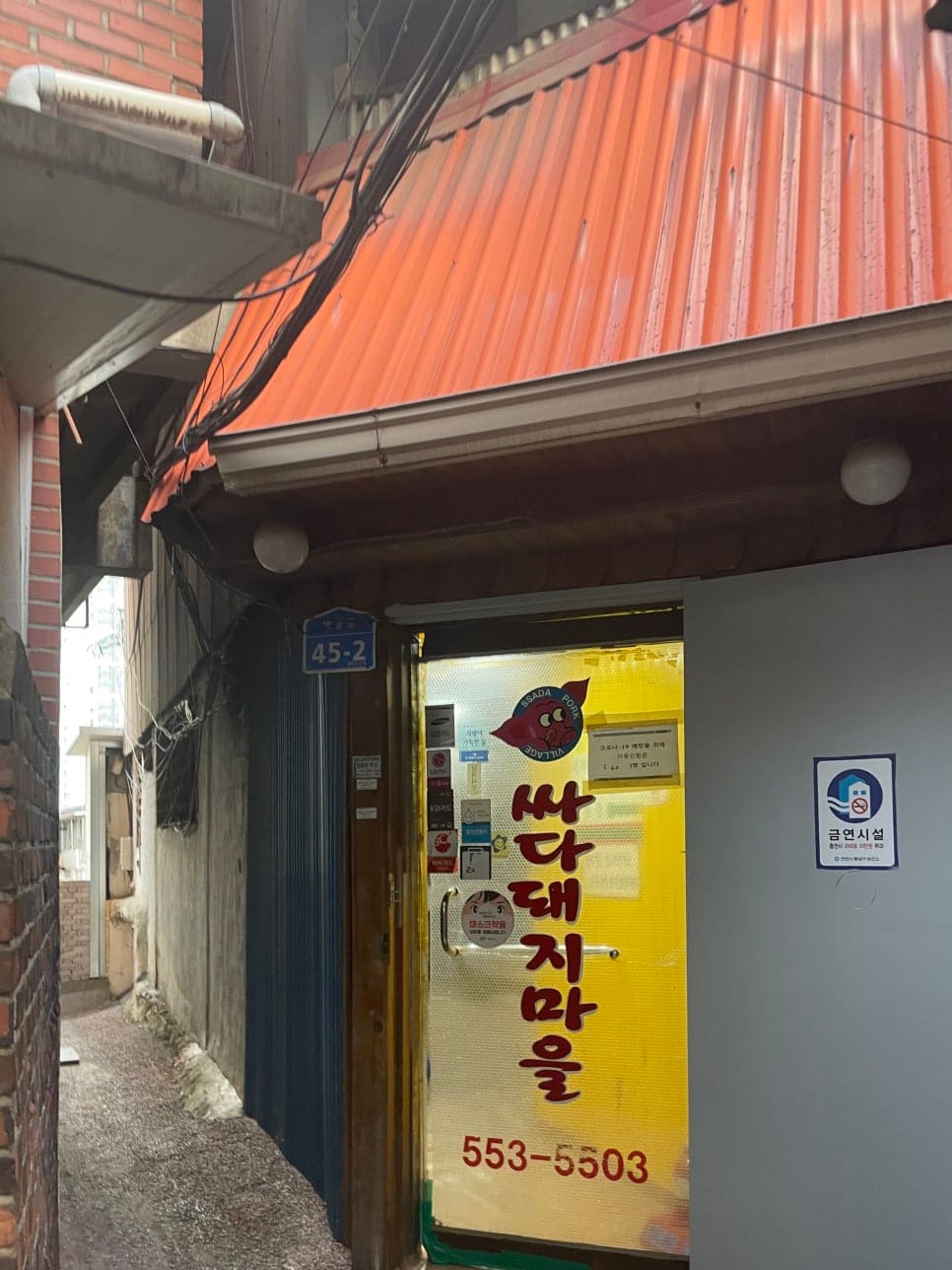 myj1112가 싸다돼지마을로 들어가는 골목 입구를 찍은 사진입니다. 주황색 함석 차양 아래 붉은 벽돌 건물이 이어지고, 번지수 표지판 45-2가 붙어 있습니다.