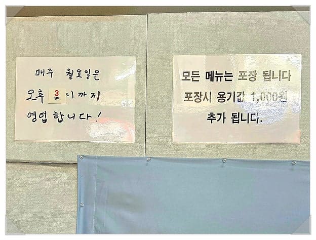 벽에 붙은 영업시간 안내문입니다. 매주 월요일은 오후 3시까지만 영업한다는 손글씨와, 모든 메뉴가 포장되고 용기값 1,000원이 추가된다는 안내가 적혀 있습니다.