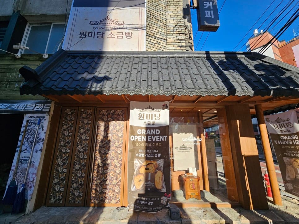 원미당소금빵 가게 앞에서 찍은 사진입니다. 기와지붕을 얹은 팝업 매장이 전선이 어지럽게 늘어진 간판 거리 한복판에 자리를 잡은 모습으로,