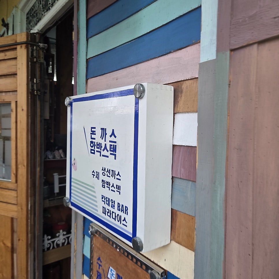 laura_oh7이 찍은 사진입니다. 돈가스·함박스테이크 메뉴가 적힌 노란 안내판과 내려진 파란 셔터, 그리고 알록달록 페인트가 칠해진 벽과 열린 출입구를 함께 담았습니다.