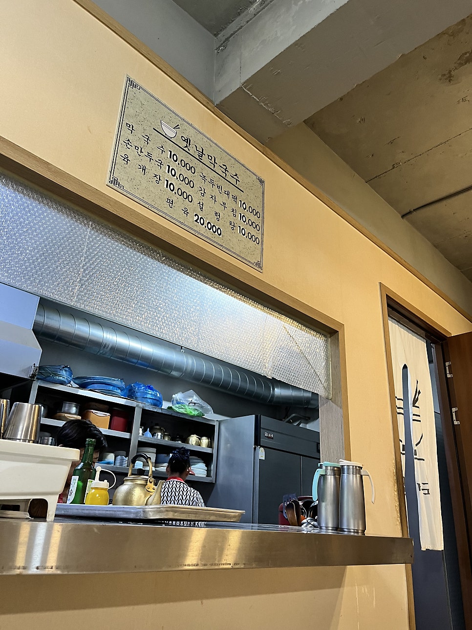 sejings가 찍은 사진입니다. 콘크리트 건물 2층으로 오르는 입구에 걸린 하얀 간판과 화분들, 그리고 안쪽 주방 위에 걸린 메뉴 가격표까지 담았습니다.