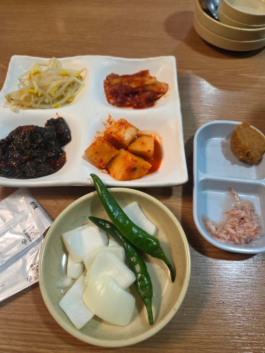 벽에 걸린 메뉴판에는 모듬국밥부터 막창전골까지 가격이 빼곡히 적혀 있고, 그 아래 '매주 화요일은 쉽니다'라는 안내문도 함께 붙어 있습니다. 다른 사진은 콩나물무침과 깻잎김치,