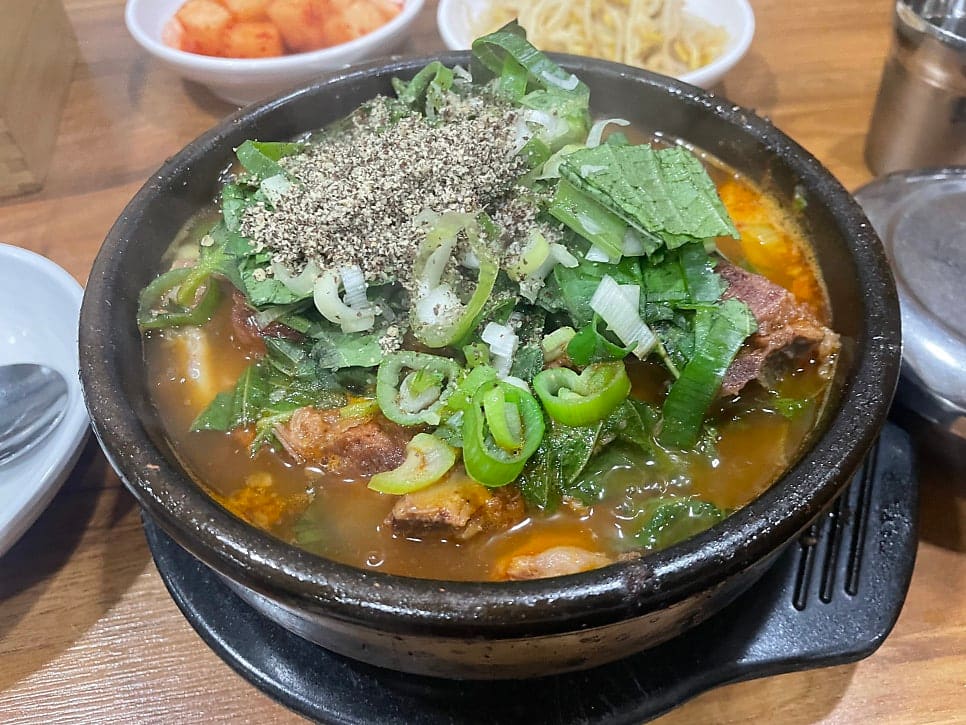 국물 요리가 담긴 뚝배기. 야채와 고기가 들어간 갈색 국물 위에 초록색 파와 참깨가 뿌려져 있다.