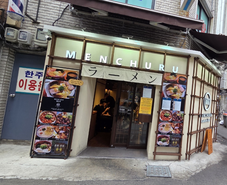 mseewon은 격자 유리창 안쪽 로고와 'MENCHURU Ramen bar' 글자, 그 아래 '해장라멘전문점'이라 적힌 나무 팻말을 가까이서 찍었습니다.