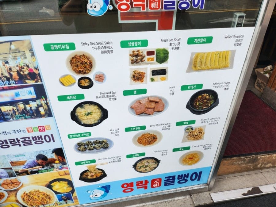 2andc는 외벽에 붙은 메뉴판과 나무 기둥에 걸린 가격표를 찍었습니다. 골뱅이무침과 생골뱅이, 계란말이 같은 메뉴 이름 아래 영어와 일본어 표기가 함께 적혀 있고,