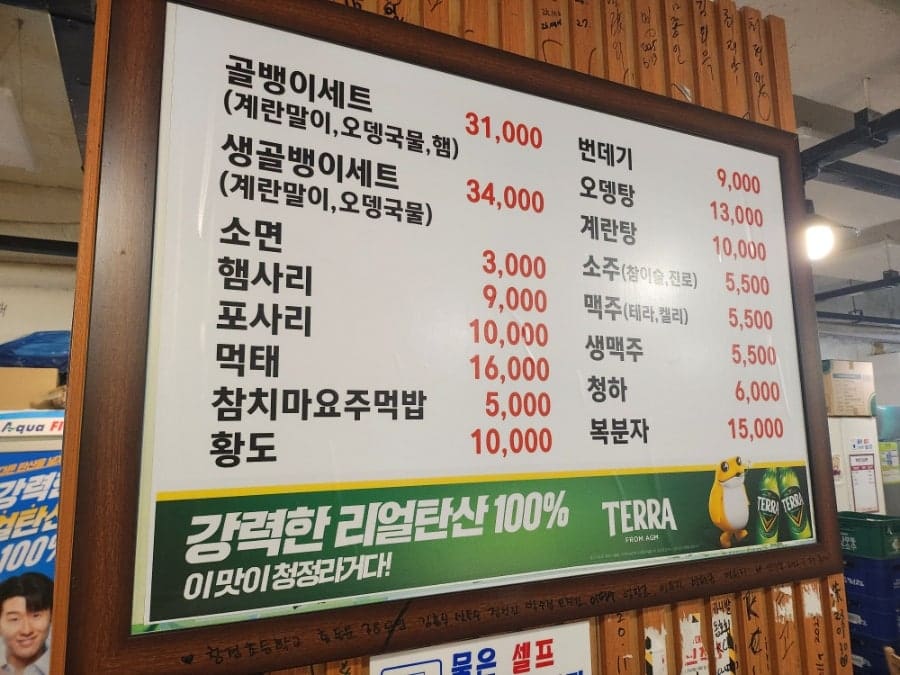 2andc는 외벽에 붙은 메뉴판과 나무 기둥에 걸린 가격표를 찍었습니다. 골뱅이무침과 생골뱅이, 계란말이 같은 메뉴 이름 아래 영어와 일본어 표기가 함께 적혀 있고,