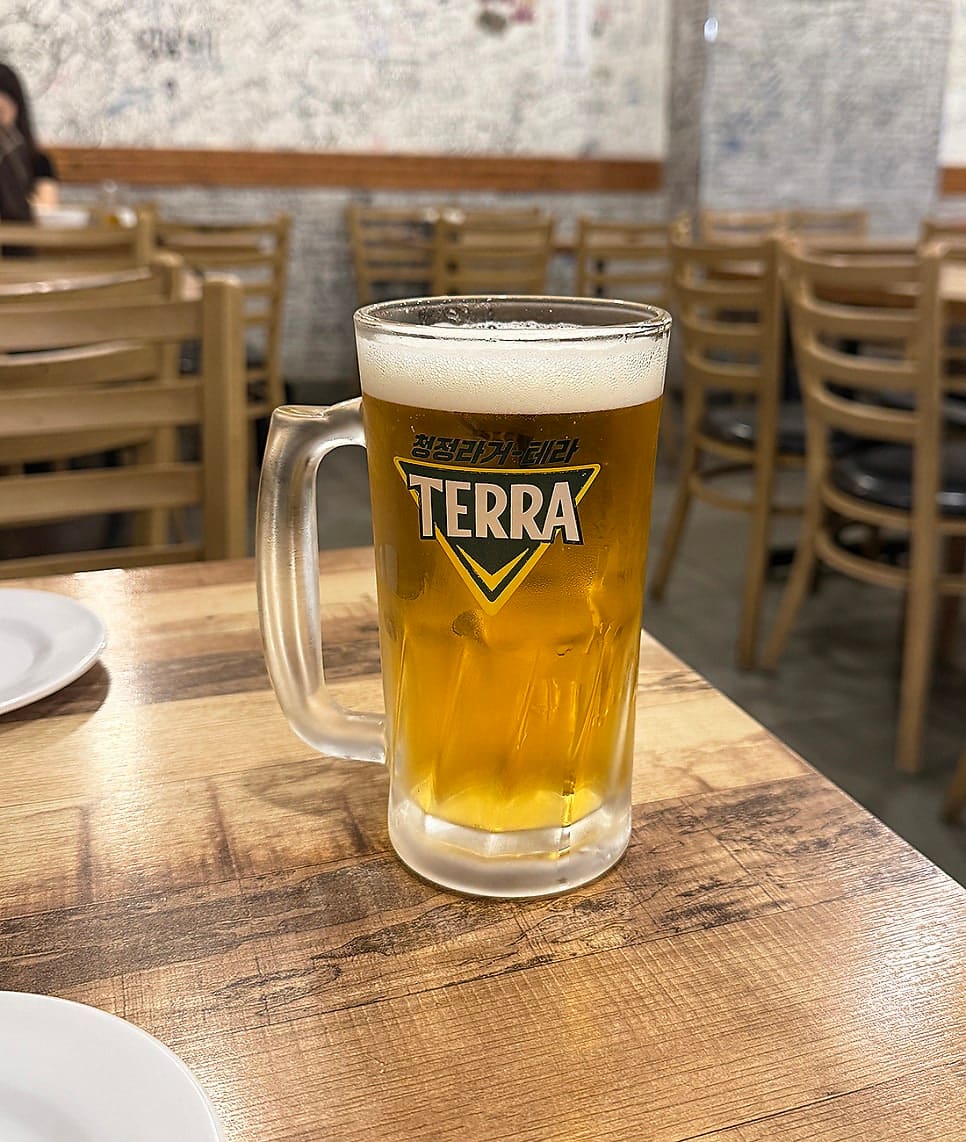 mmmmhhzz는 자리에 앉자마자 시킨 생맥주를 찍었습니다. 김이 서린 잔에 거품이 두툼하게 얹혀 있고, 뒤로는 빈 테이블과 의자가 줄지어 보입니다.