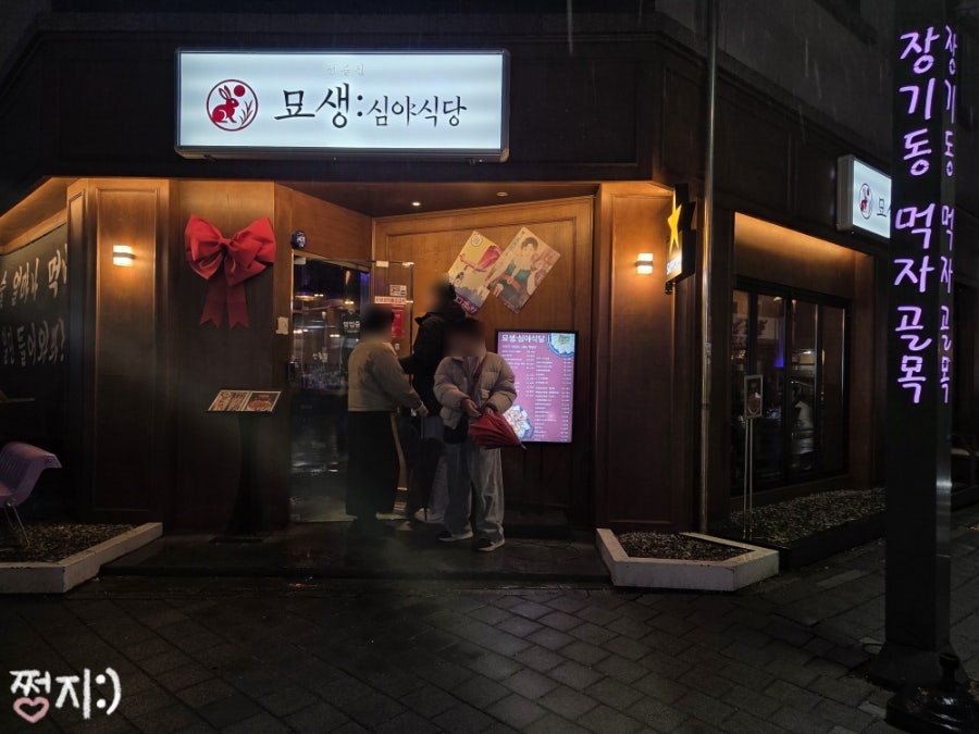 wus7845는 비 오는 밤 우산을 쓰고 줄 서 있는 일행과, 옆 건물의 '장기동 먹자골목' 세로 네온 간판을 함께 담았습니다.