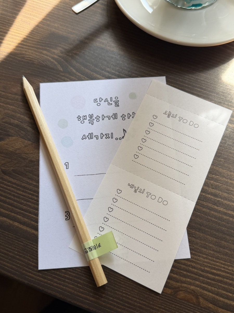 같은 테이블을 가까이서 담은 사진입니다. 나를 행복하게 하는 세 가지를 적어보라는 메모지와 오늘의 할 일 체크리스트가 나란히 놓여,