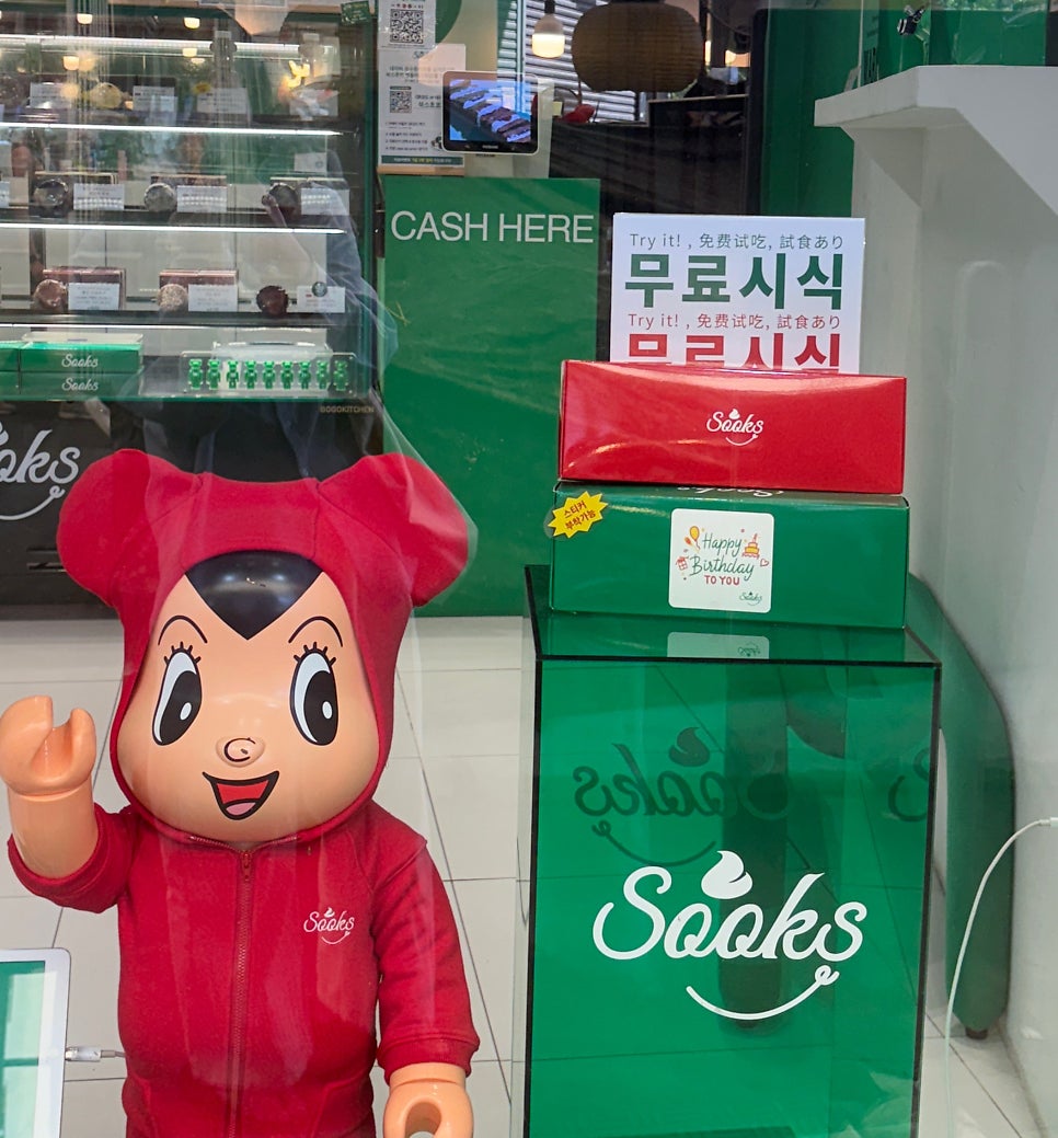 빨간 후드를 쓴 마스코트 인형 옆으로 '무료시식' 안내문과 생일 축하 스티커가 붙은 선물 상자가 쌓여 있습니다.