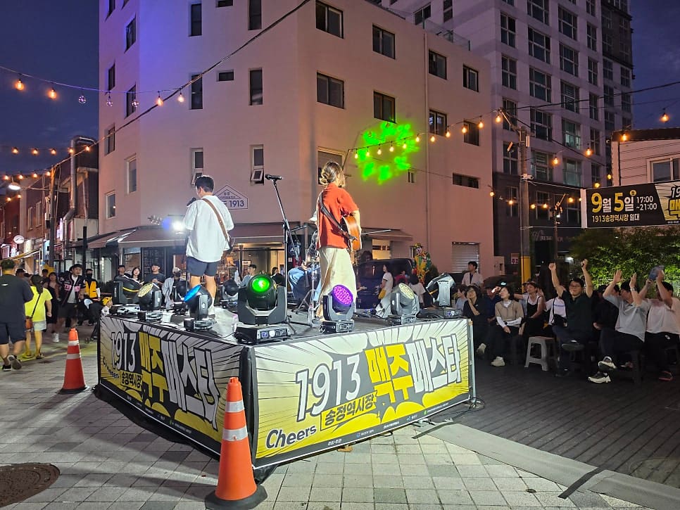 amawin님이 맥주페스타가 열린 금요일 밤 남긴 사진입니다. 알전구 조명 아래 무대 위에서 버스커가 노래하고, 플라스틱 의자에 둘러앉은 사람들이 맥주잔을 들고 있습니다.