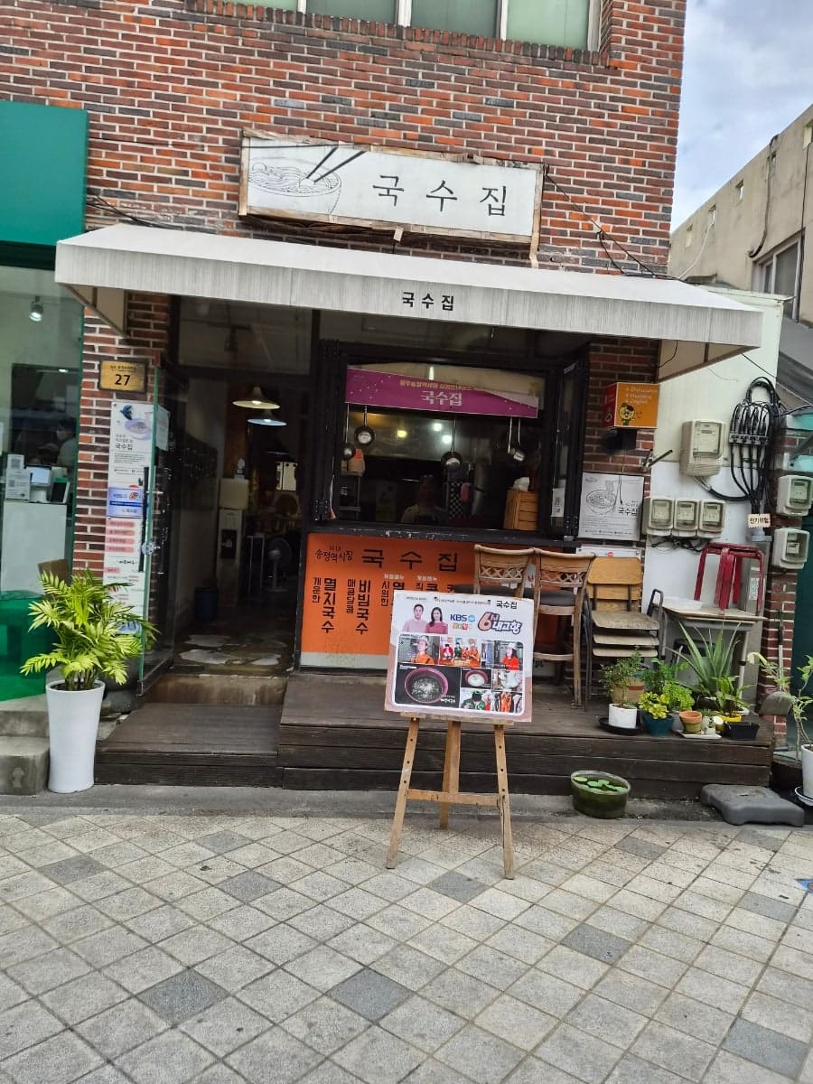 languagebuddy님이 시장 골목을 걸으며 남긴 사진입니다. 한쪽에는 손글씨 메뉴판을 내건 오래된 국수집 벽에 지역 방송 출연을 알리는 안내판이 붙어 있고,