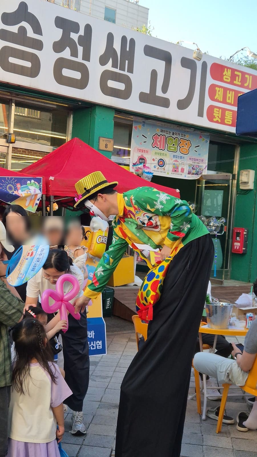 start0347님이 맥주페스타 현장에서 남긴 사진입니다. 삐에로 복장을 한 공연자가 풍선을 만들어 아이들에게 건네고, 뒤로는 오래된 정육점 간판과 체험장 현수막이 함께 보입니다.