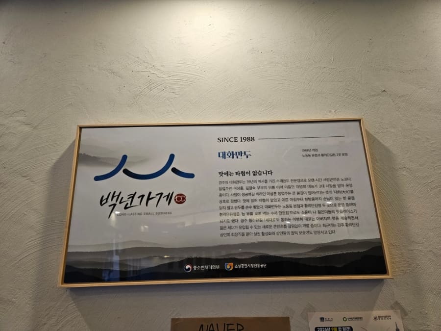 midali1981이 담은 입구와 벽면입니다. 나무 벤치가 놓인 오픈형 매장 입구와, '맛에는 타협이 없습니다'라는 문구가 적힌 백년가게 인증 명패가 함께 찍혔습니다.