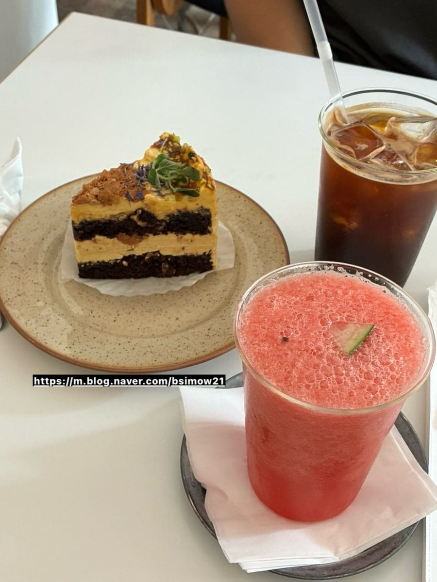 bsimow21님이 남긴 사진입니다. 백합과 도자기 소품이 놓인 선반 너머로 만석인 홀이 보이고, 앞쪽 테이블에는 피넛버터초코케이크와 수박주스, 아메리카노가 놓여 있습니다.