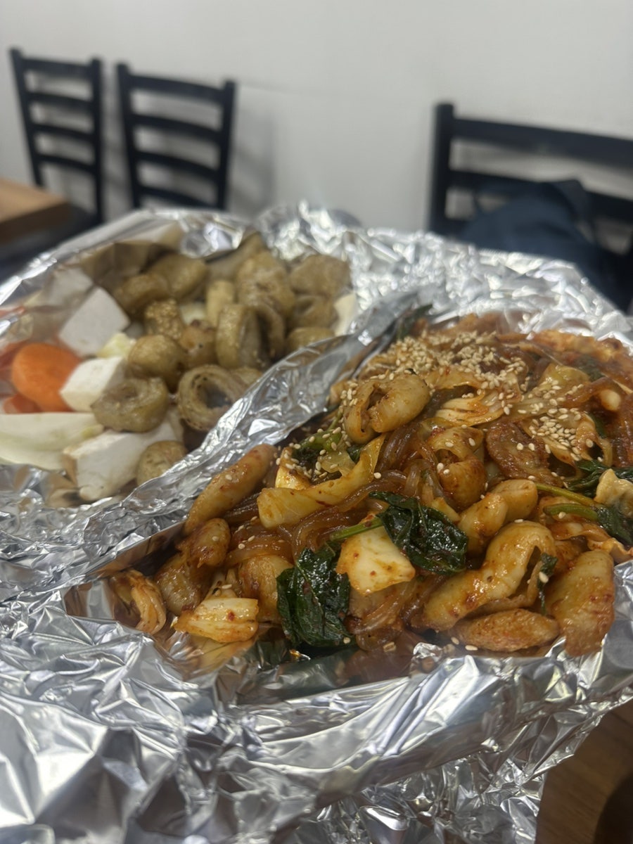 travel_me가 촬영한 상차림과 포일에 담긴 야채곱창입니다. 스테인리스 컵과 쌈채소 바구니가 놓인 테이블 위로,