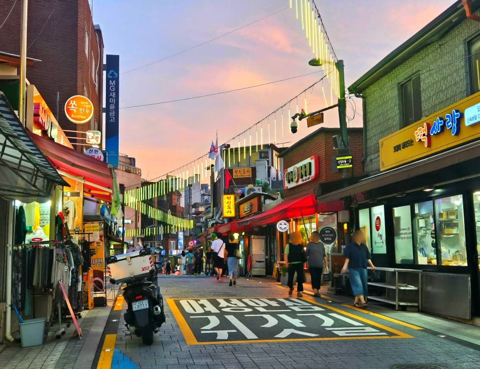 jhbluesss가 해 질 녘에 촬영한 골목 풍경입니다. 색색의 전구 줄과 네온 간판이 하나둘 켜지며, 낮 동안 조용하던 거리가 사람들의 말소리로 채워지기 시작합니다.