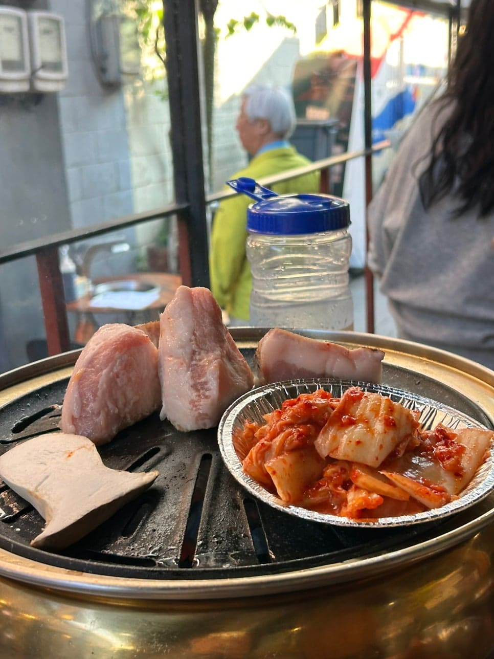 7bae9lee6은 야외 화로 앞에 앉아, 아직 굽기 전인 고기와 김치가 놓인 상 너머로 골목을 지나는 사람들을 함께 담았습니다.
