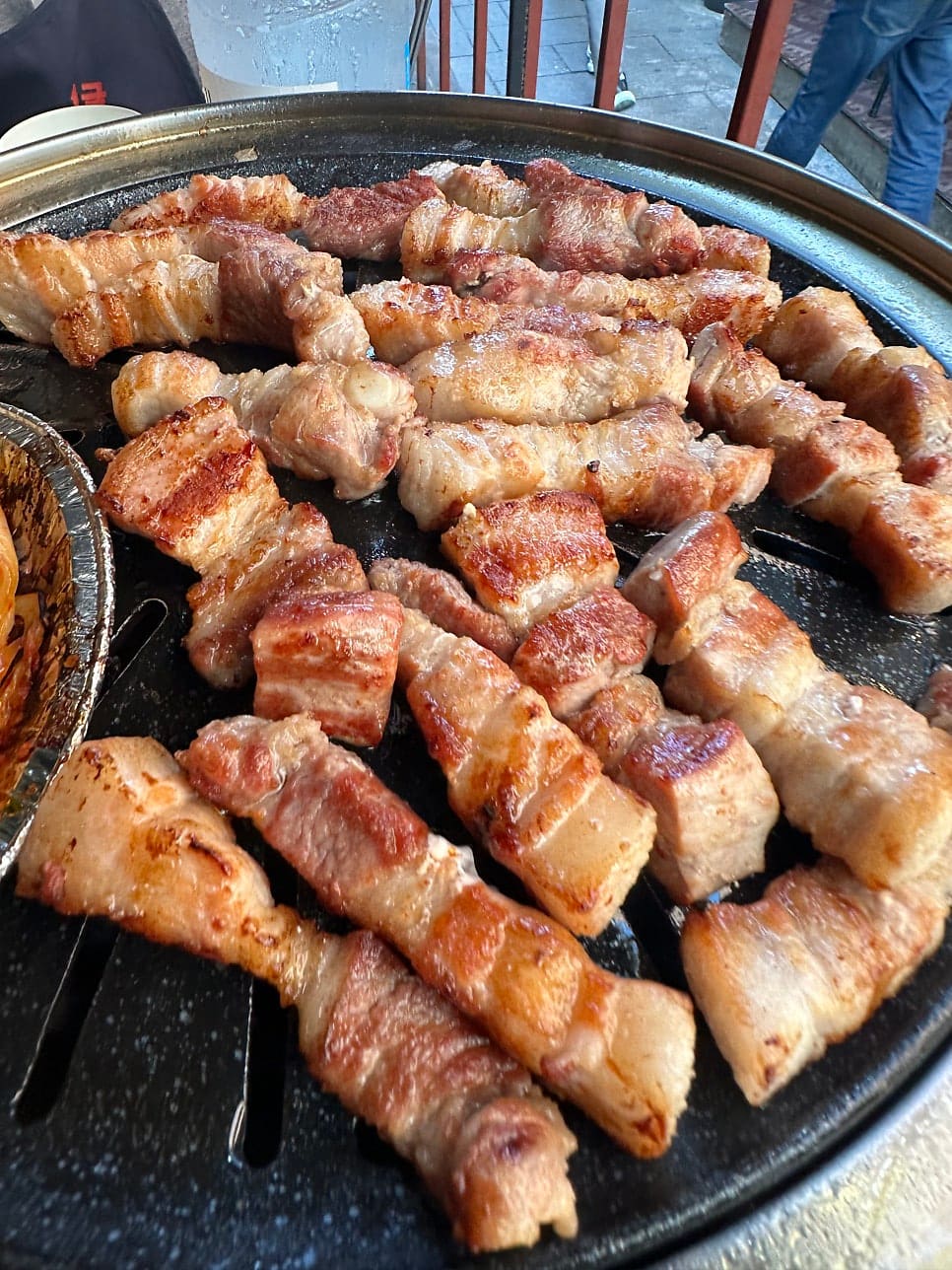 7bae9lee6은 야외 화로 앞에 앉아, 아직 굽기 전인 고기와 김치가 놓인 상 너머로 골목을 지나는 사람들을 함께 담았습니다.