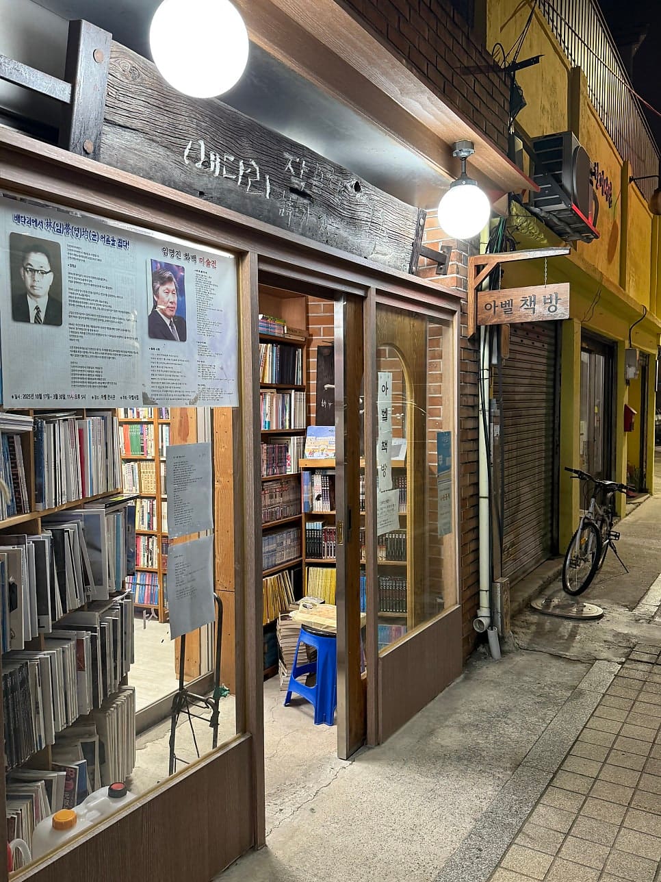 저녁이 내려앉은 골목, 집현전과 아벨책방의 불빛이 좁은 인도를 비춘다. 자전거 한 대와 화분들이 문 앞을 지키고 있다.