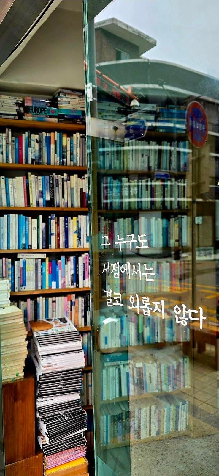 책방 유리문에 붙은 손글씨, 그 누구도 서점에서는 결코 외롭지 않다. 옆 책방 서가에는 거리 풍경이 유리에 그대로 겹쳐 비친다.